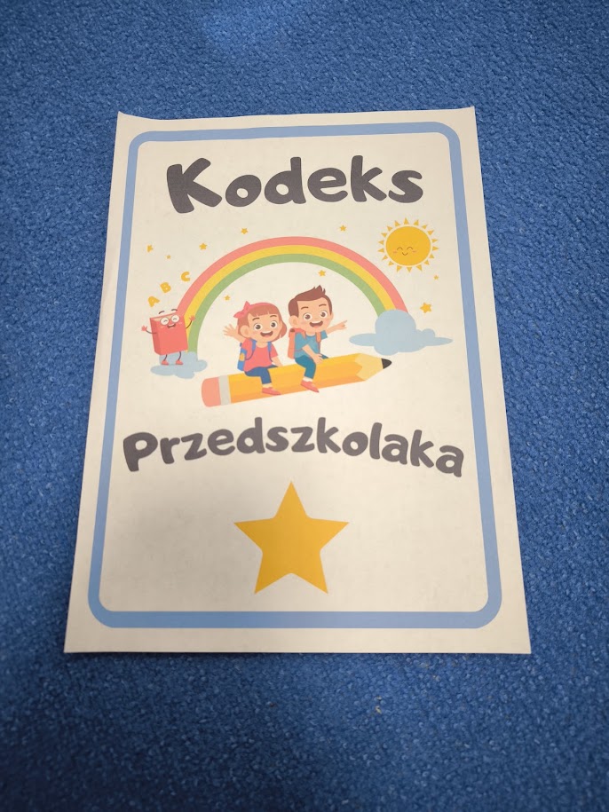 Okładka „Kodeks Przedszkolaka” z tęczą, dwojgiem dzieci na ołówku i żółtą gwiazdką, leżąca na niebieskim dywanie