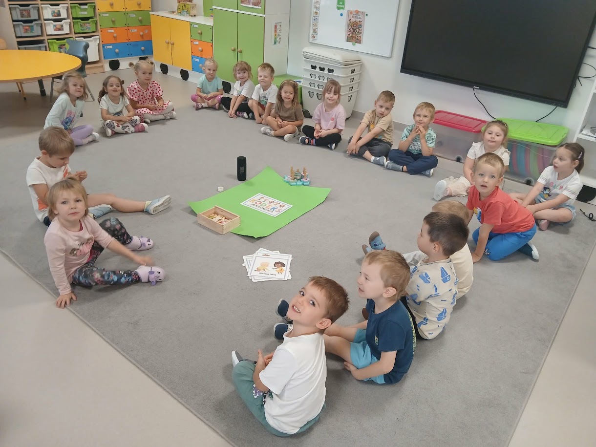 Dzieci siedzą w kręgu na dywanie w sali; na środku zielony arkusz i obrazki do ustalania kodeksu grupowego